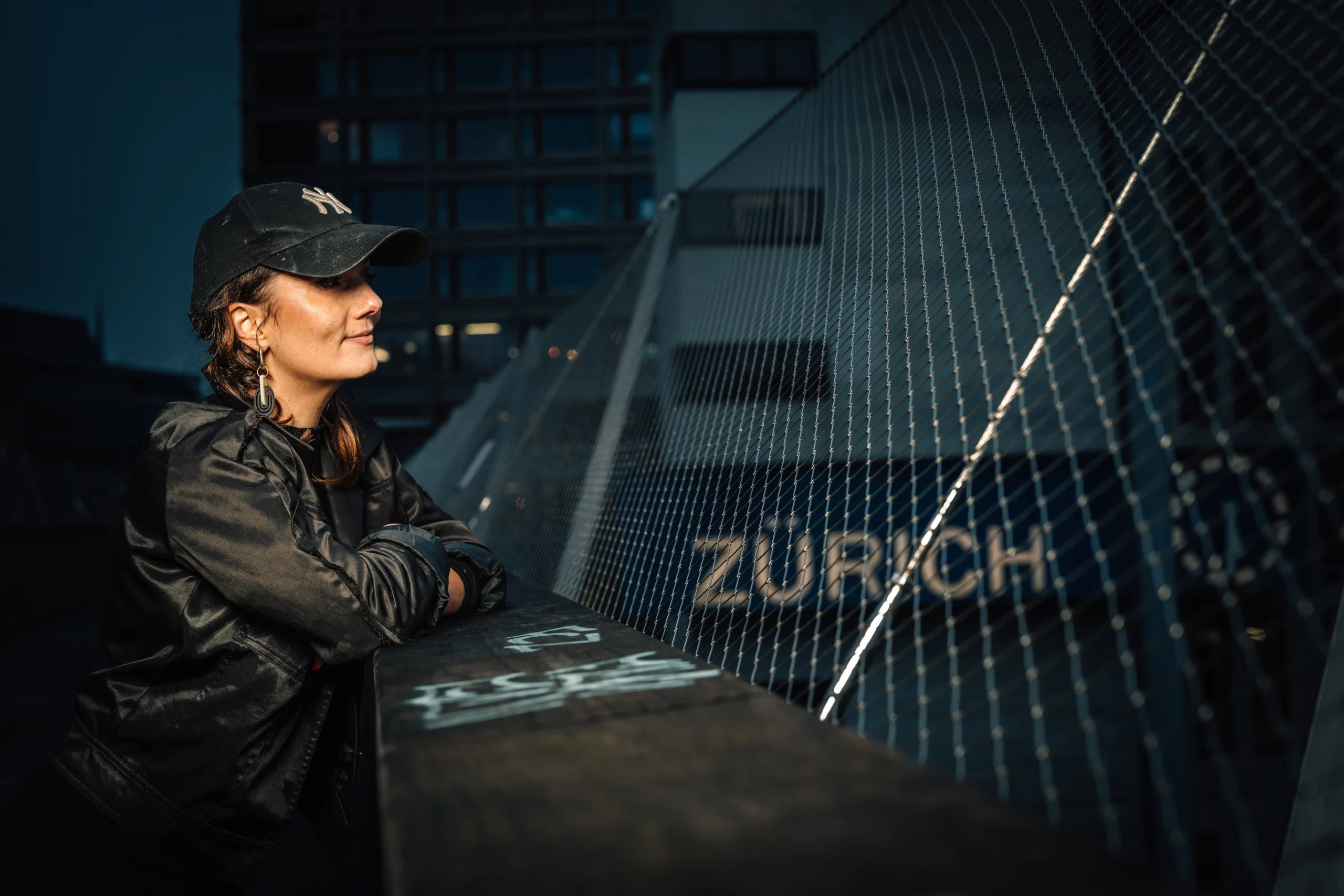 Rosanna Grüter lehnt sich auf ein Brückengelender und schaut nach rechts in die Ferne, mit dem Schriftzug des Bahnhof Zürich im Hintergrund sichtbar.